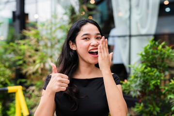 Young Asian woman in black top covering mouth as if checking breath, giving a satisfied thumbs up, in approval of her breath, outside a blurred cafe setting. background.