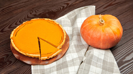 Pumpkin pie on a wooden table, with a slice removed, accompanied by a fresh pumpkin and a checkered...
