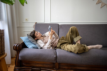 Procrastinating young woman lounging on sofa browsing chat apps on smartphone, laziness and...