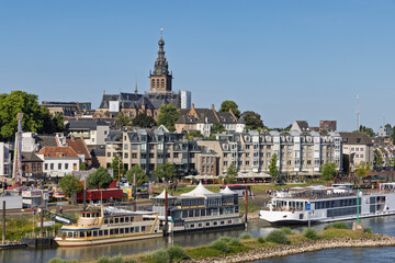 Fototapeta premium The historic city of Nijmegen, Netherlands, seen from the riverside with the prominent St. Stephen's Church and boats docked along the quay. Nijmegen, Netherlands. 20 June 2025.