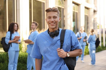 Happy medical student in uniform with backpack and headphones near building outdoors, selective focus
