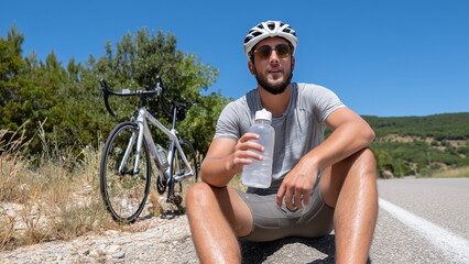 A biker with helmet on resting on the roadside, drinking water after an intense ride, sweat glistening, capturing a moment of recovery and endurance in an outdoor cycling setting.