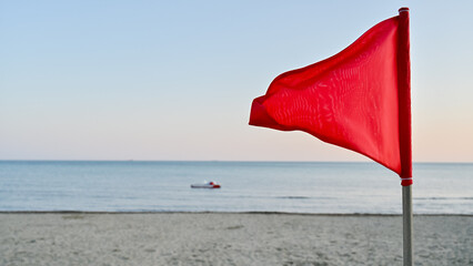 Out-of-focus red triangular warning flag in the foreground, with a sharp focus on the calm but potentially dangerous sea in the background. A pedal boat floats near the horizon.