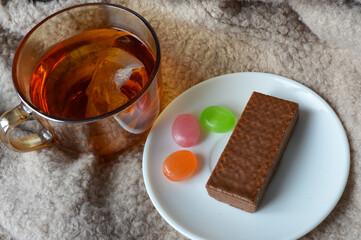 glass cup of black tea with a chocolate wafer and colorful hard candies on a white plate placed on...