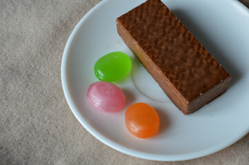 glass cup of black tea with a chocolate wafer and colorful hard candies on a white plate placed on soft beige fabric tea time aesthetics, cozy moments, dessert and confectionery, relaxation fall