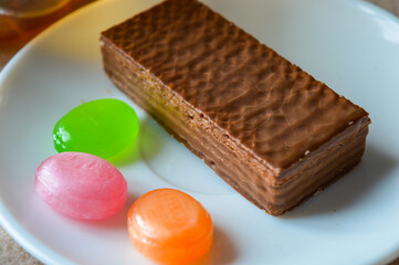 glass cup of black tea with a chocolate wafer and colorful hard candies on a white plate placed on soft beige fabric tea time aesthetics, cozy moments, dessert and confectionery, relaxation fall