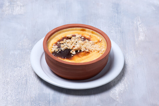 High angle studio photo of baked milk pudding with crcushed hazelnut topping  in earthenware pot, a traditonal Turkish dessert called fırında s&uuml;tla&ccedil; on white wooden table