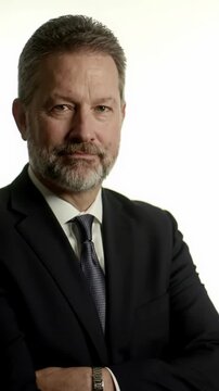 A confident mature man in a dark business suit with a salt-and-pepper beard maintains eye contact, smiling subtly then shifting posture on a brightly lit pure white studio background, conveying