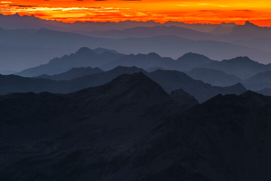 Layered mountain ranges fade into the distance under a vibrant orange and red sunset. Mantova refuge al Vioz, Peio, Trentino. The sky is streaked with colorful clouds.