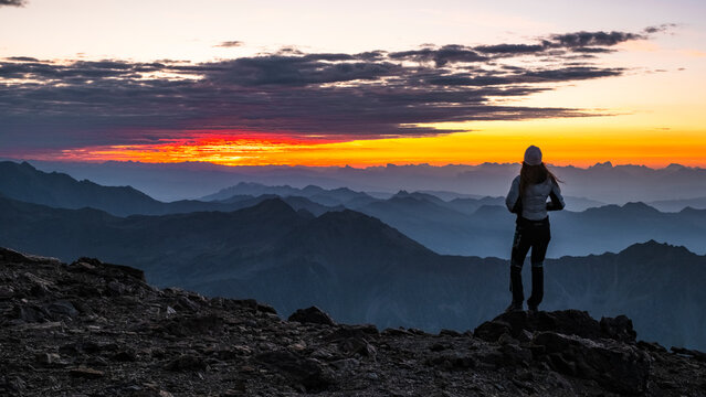 A woman stands on a rocky peak, silhouetted against a vibrant sunrise over misty mountain ranges. The sky transitions from dark clouds to orange and yellow hues. Mantova refuge al Vioz, Peio,
