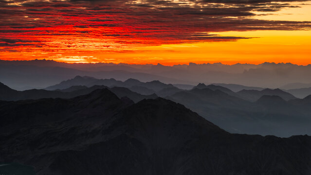 A landscape of layered mountain ranges silhouetted against a vibrant orange and red sunset. Mantova refuge al Vioz, Peio, Trentino. 