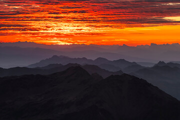 A vibrant orange sunset over layered mountain ranges. The sky is filled with textured clouds. Mantova refuge al Vioz, Peio, Trentino. The peaks are silhouetted against the bright sky.