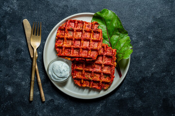 Vegetarian beetroot and zucchini waffles with herbs on a plate with sour cream on table, closeup, top view. Tasty vegetable waffles for eat made with waffle maker