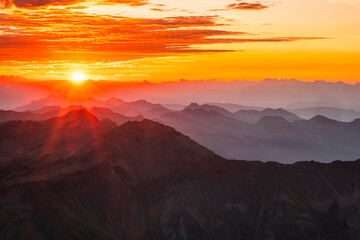 Sunrise over misty mountain ranges, with orange sky and clouds. Mantova refuge al Vioz, Peio, Trentino. The sun's rays illuminate the peaks in the foreground.