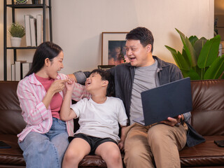 A father, mother and son sit on the sofa in the living room. The father holds a laptop and invites them to watch.