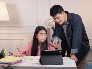 Asian dad helps his daughter do her homework at the table with a tablet.