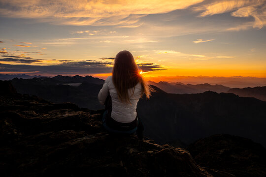 A woman sits on a rocky peak, back to the camera, watching a vibrant sunset over distant mountains. The sun shines through her hair. Mantova refuge al Vioz, Peio, Trentino.