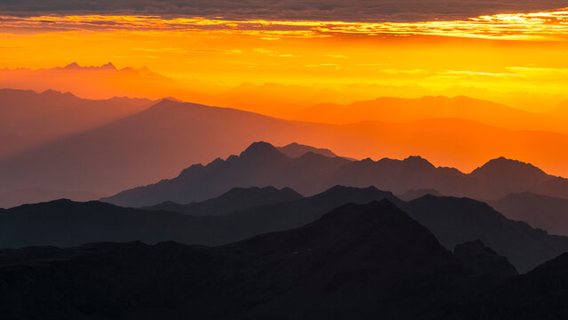 Layers of dark mountain silhouettes fade into an orange sky at sunset. The sky is bright orange with some clouds at the top. Mantova refuge al Vioz, Peio, Trentino 