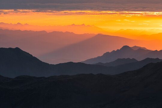 Layers of dark mountain silhouettes fade into an orange sky at sunset. The sky is bright orange with some clouds at the top. Mantova refuge al Vioz, Peio, Trentino 