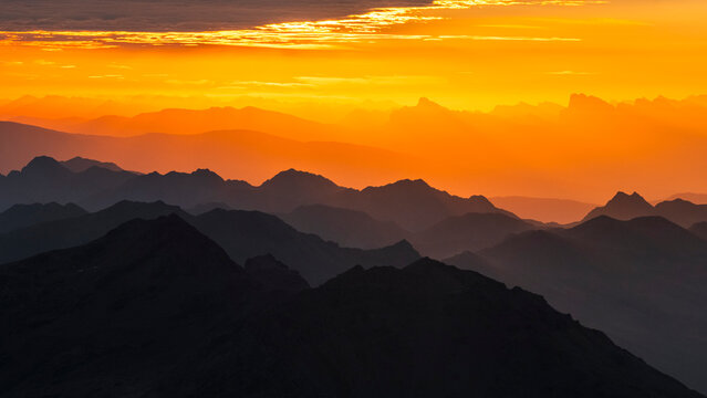 Layers of dark mountain silhouettes fade into an orange sky at sunset. The sky is bright orange with some clouds at the top. Mantova refuge al Vioz, Peio, Trentino 