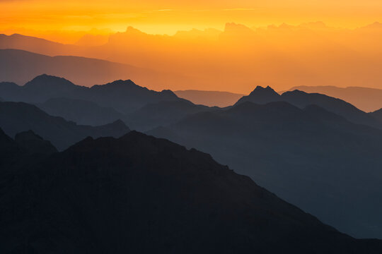Layers of dark mountain silhouettes fade into an orange sky at sunset. The sky is bright orange with some clouds at the top. Mantova refuge al Vioz, Peio, Trentino 