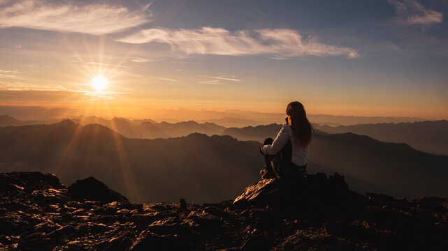 A woman sits on a rocky peak, silhouetted against a sunrise over distant mountains. The sun bursts through the clouds, casting rays across the landscape. Mantova refuge al Vioz, Peio, Trentino.