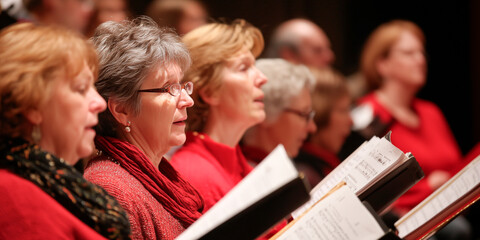A group of choir members dressed in red passionately singing together, holding sheet music, creating an atmosphere of joy and harmony during a festive performance.

