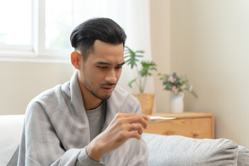 Seasonal cold sickness in people concept, Sick asian man looking at thermometer during measuring...