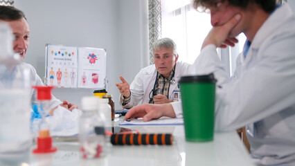The chief of surgery at the meeting talks about the clinic's plans. Three doctors are sitting at a table in a meeting room, with a poster of a human body