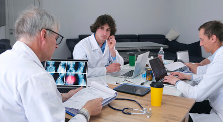 A group of doctors are sitting at a table with laptops and discussing laboratory tests. The head physician of the hospital advises his colleagues on surgery issues
