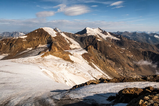 A high-angle shot of snow-capped mountains under a blue sky with scattered clouds. The foreground shows a rocky, snowy terrain. View from the top  of Vioz Mount, Trentino.