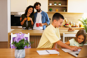 Happy family enjoying morning routine in kitchen: parents drinking coffee while children doing homework