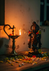 A small statue of a Hindu deity is illuminated by the soft glow of a traditional oil lamp, surrounded by flower petals on a wooden surface