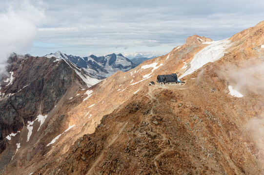 Mantova refuge al Vioz, Peio, Trentino on a rocky slope with snow patches, against a backdrop of distant peaks and cloudy sky. The refuge is a dark building with a light stone base.