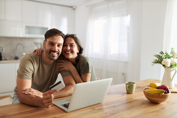 Happy couple using credit card for online shopping over laptop at home and looking at camera.
