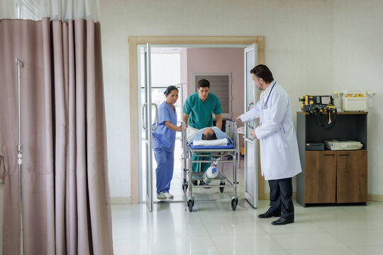 Medical team moving injured asian male patient on stretcher into hospital emergency room while senior caucasian male doctor standing beside door preparing to assist during urgent care process
