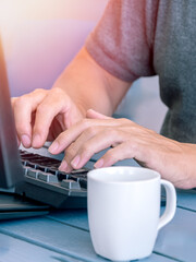Hands typing on keyboard with coffee cup nearby 2.- Close-up of hands typing on a keyboard with a white coffee cup on a blue table.
