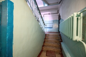 stairway with faded, pastel-colored walls leads upwards. The worn stairs and a painted radiator give a sense of age and utilitarian function