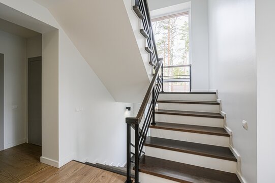 modern staircase with wooden steps and white risers ascends towards a window with trees outside. The railing is dark metal. The walls are white, and the floor is light wood