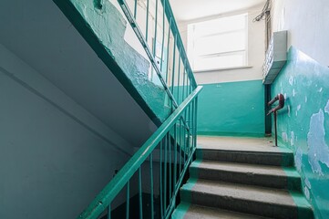 stairwell with green handrails and blue walls, showing signs of age and wear. A window is visible...