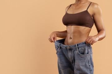 Cropped image of toned woman in brown underwear holding oversized jeans. Symbol of significant weight loss, fitness goal achievement, and healthy lifestyle transformation, copy space