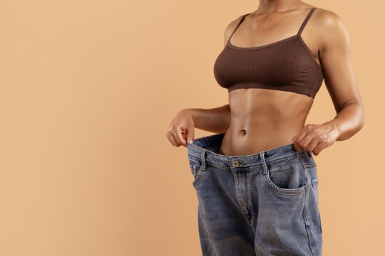 Cropped image of toned woman in brown underwear holding oversized jeans. Symbol of significant weight loss, fitness goal achievement, and healthy lifestyle transformation, copy space - Powered by Adobe