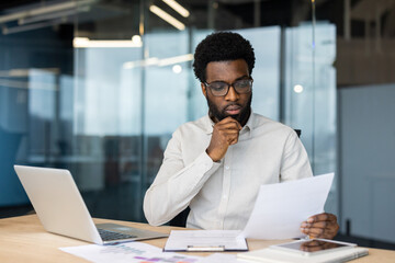 Obraz premium A businessman attentively examines paperwork at his desk in a contemporary office. He is surrounded by modern tools, including a laptop and smartphone, highlighting a setting of productivity