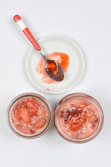 A top-down view of two jars of homemade strawberry jam and a small plate with a tasting spoon on a gray background