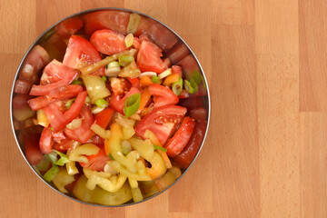 A vibrant tomato and pepper salad in a metal bowl, placed on a light wooden background with ample copy space on the right side