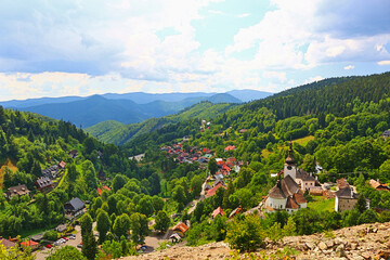 A picturesque view of a small village of Špania Dolina in Slovakia nestled in a lush green valley, surrounded by rolling hills and forests.