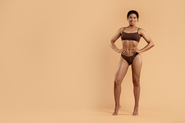 Fototapeta premium Fit African American woman in brown underwear standing with hands on waist, smiling proudly. Beige backdrop and full body shot emphasize wellness and body positivity, copy space