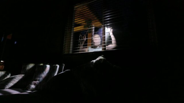 masked man holding a crowbar and looking into a home through window blinds at night. The unsettling atmosphere evokes fear and suspense, ideal for illustrating concepts of crime, danger, home invasion