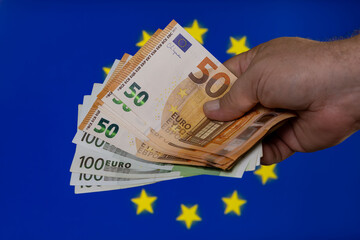 Hand holds several euro banknotes against the backdrop of the European Union flag, showcasing financial currency in a relevant context