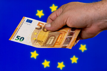 Person holding a fifty euro banknote in front of a blue European Union flag background during a financial transaction or commerce activity